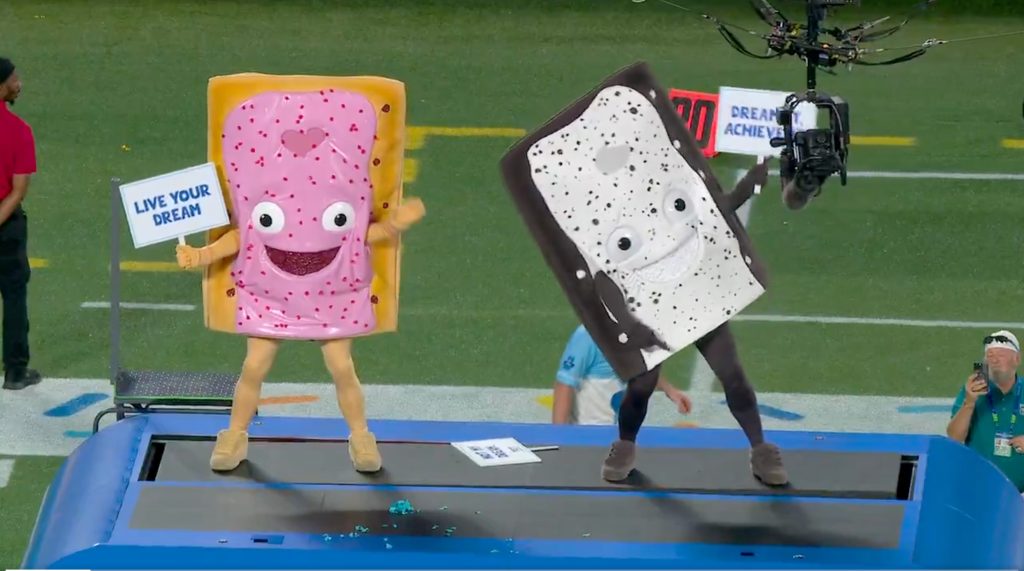 Two people in Pop-Tart mascot costumes on a platform at a football game.